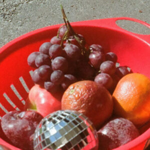 A red strainer full of grapes, apples and oranges and a small disco ball among them