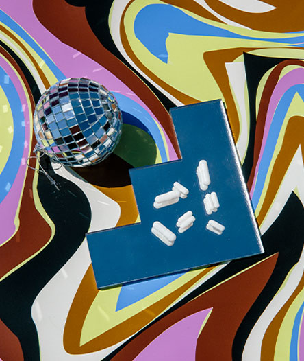 Several white pills site on a stepped mirror beside a small disco ball on a swirled colour background