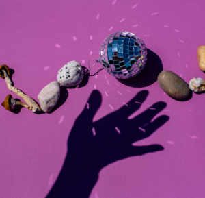 A hands shadow on a purple backdrop with rocks, a disco ball and magic mushrooms arranged in a wavy line