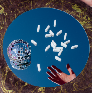 Several white pills and a disco ball sit on a mirror on the ground with a hands reflection overtop